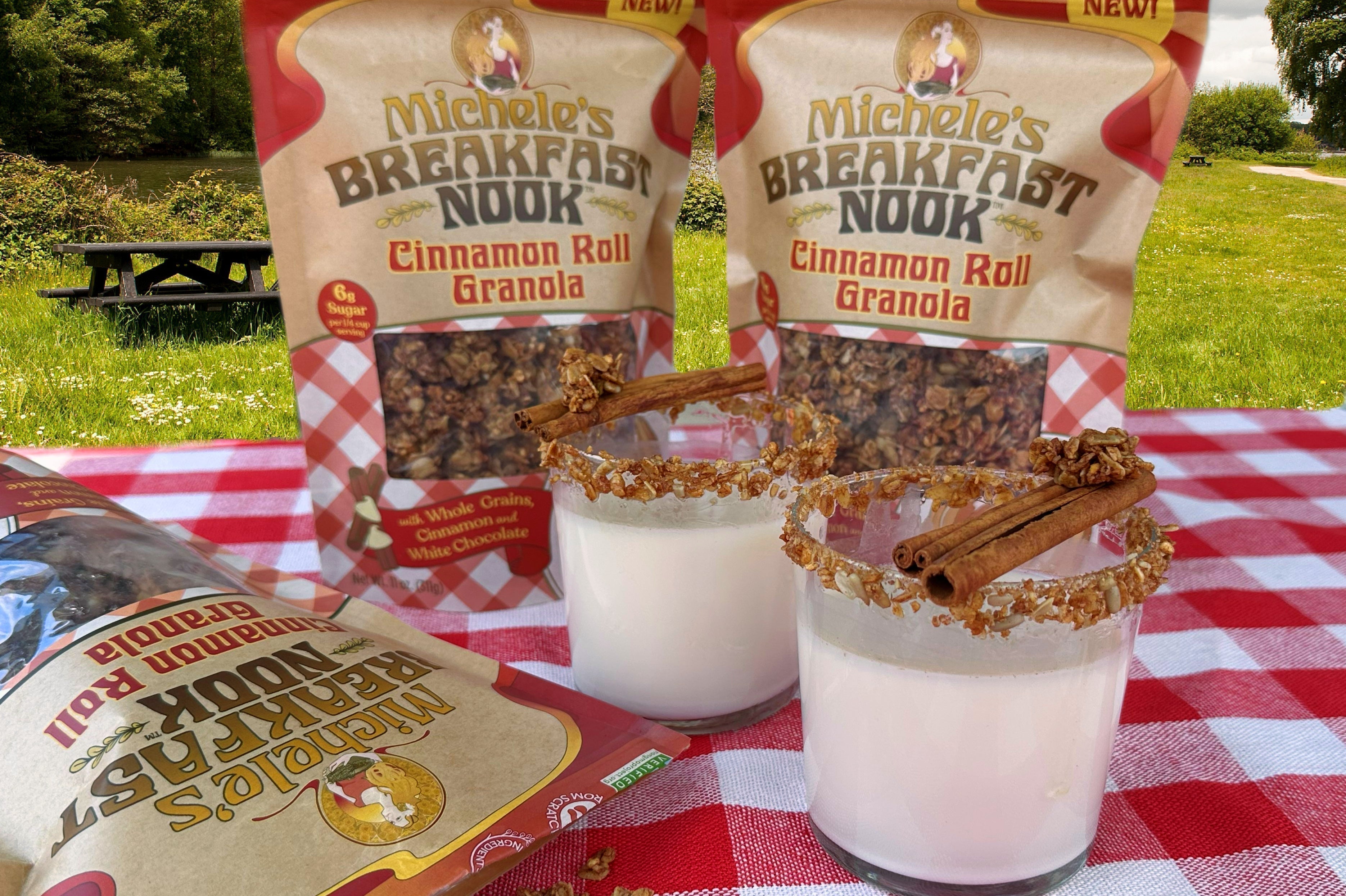 Two glasses of homemade horchata made with Michele's Breakfast Nook Cinnamon Roll Granola, garnished with granola and cinnamon sticks and served on an outdoor picnic table with a red gingham tablecloth.